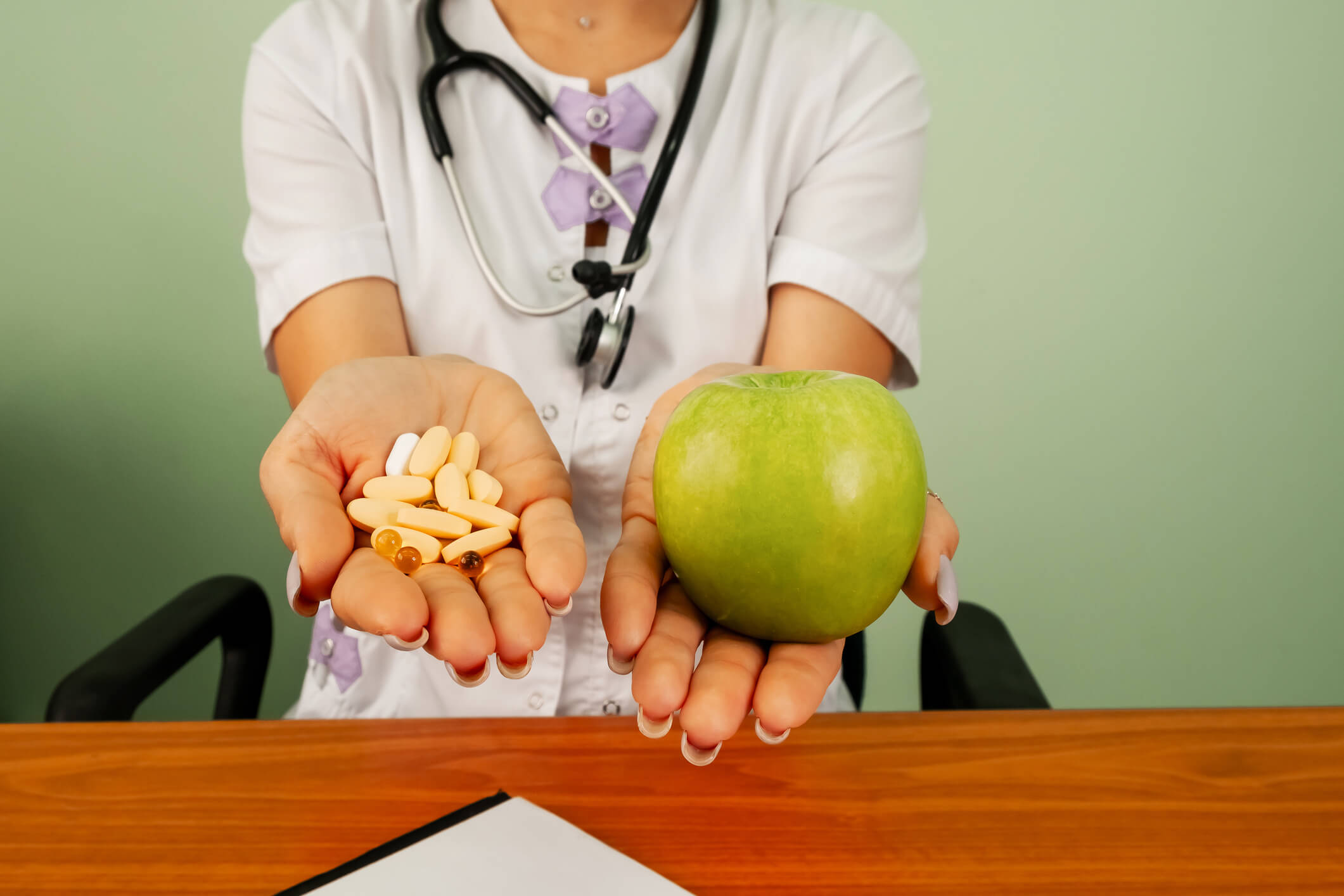 A doctor is seated before a desk. She has two outstretched hands, one of which contains some pills and the other contains an apple.