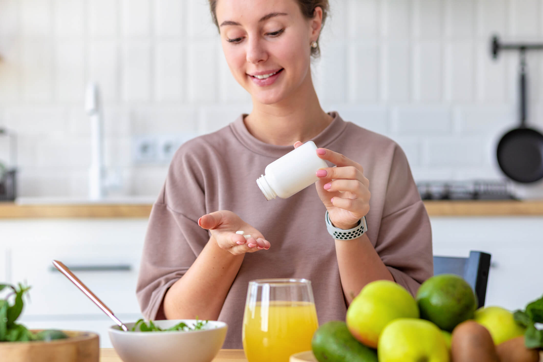 A woman has two pills in one hand and an upturned pill bottle in the other. She is seated at a table before a bowl of salad, some fruit, and a glass of juice. 