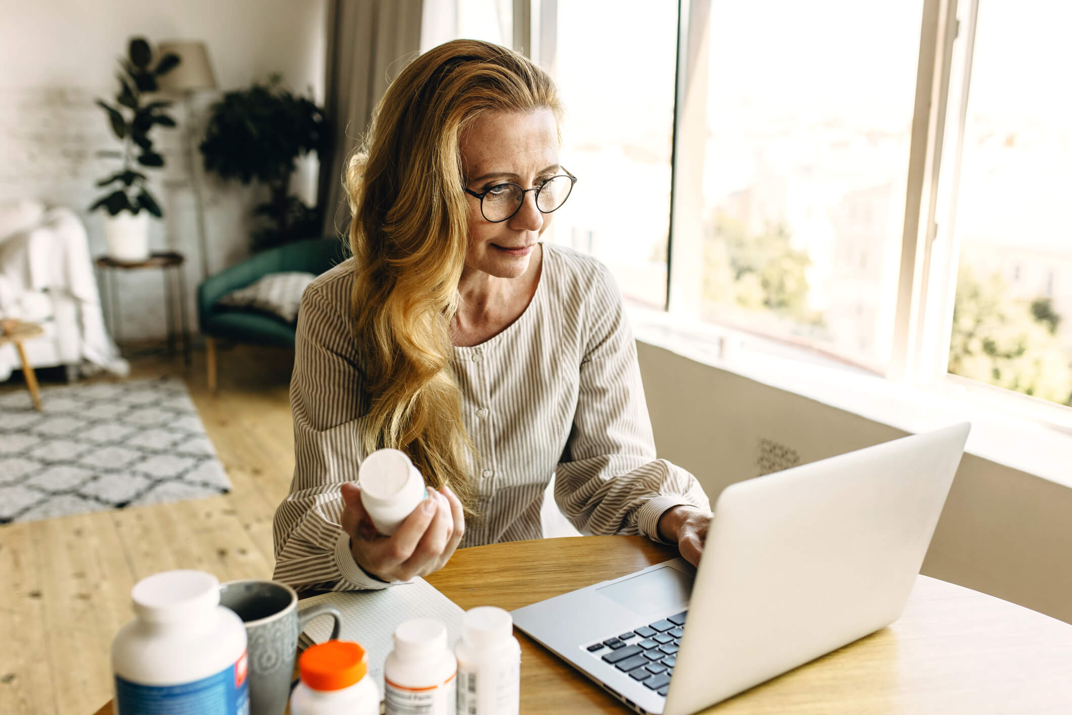 A woman is seated at a table looking at a laptop. Her left hand is on the keyboard and her right hand is holding a pill bottle that is oriented as though she is researching the text on the bottle