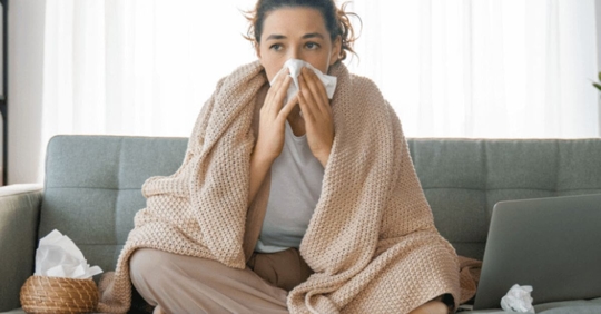 young woman sitting on couch wrapped in a blanket blowing her nose