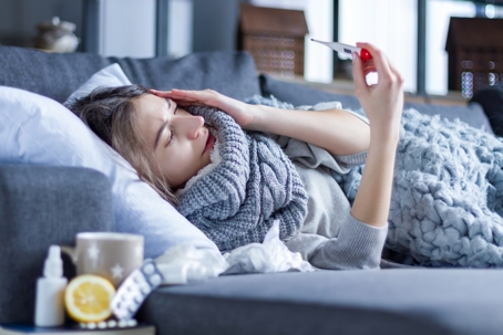 Woman with illness covered in blankets with tea and tissues next to her, holding a thermometer in hand and holding her head as she fights a fever.