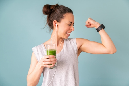 A woman holds a green drink in her right hand while flexing with her left arm, smiling.