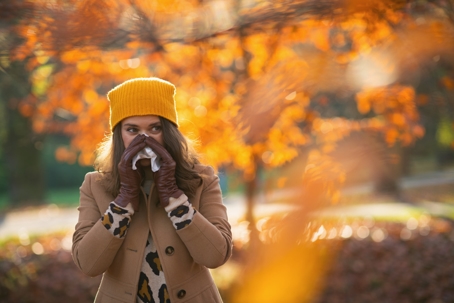 Woman with fall allergies taking a walk outside while blowing her nose. The trees behind her are changing color and she is wearing a knitted hat and jacket. | Advance ER