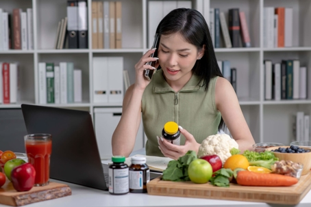 A seated woman holds a phone to here ear with her right arm, while she holds a pill in her left hand. There is a pile of vegetables and several pill bottles in front of her.