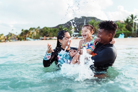 Parents on a resort vacation with their young child. They are playing in the water at the beach, laughing and splashing water around. | Advance ER