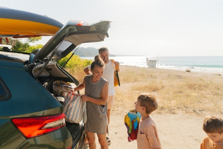 Family of four on a road trip vacation to the beach. Parents are unpacking the car while the children excitedly wait to start playing in the sand and water. | Advance ER
